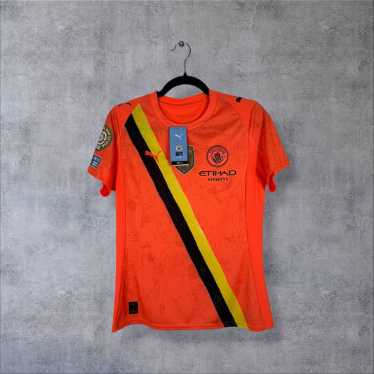 Maillot gardien Manchester City KidSuper orange de face, écharpe jaune et noire, sponsor Etihad et étiquettes attachées