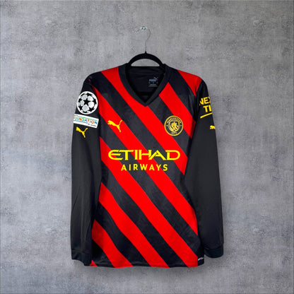 Vue de face du maillot Manchester City 2022/23 extérieur manches longues aux bandes diagonales rouges et noires.