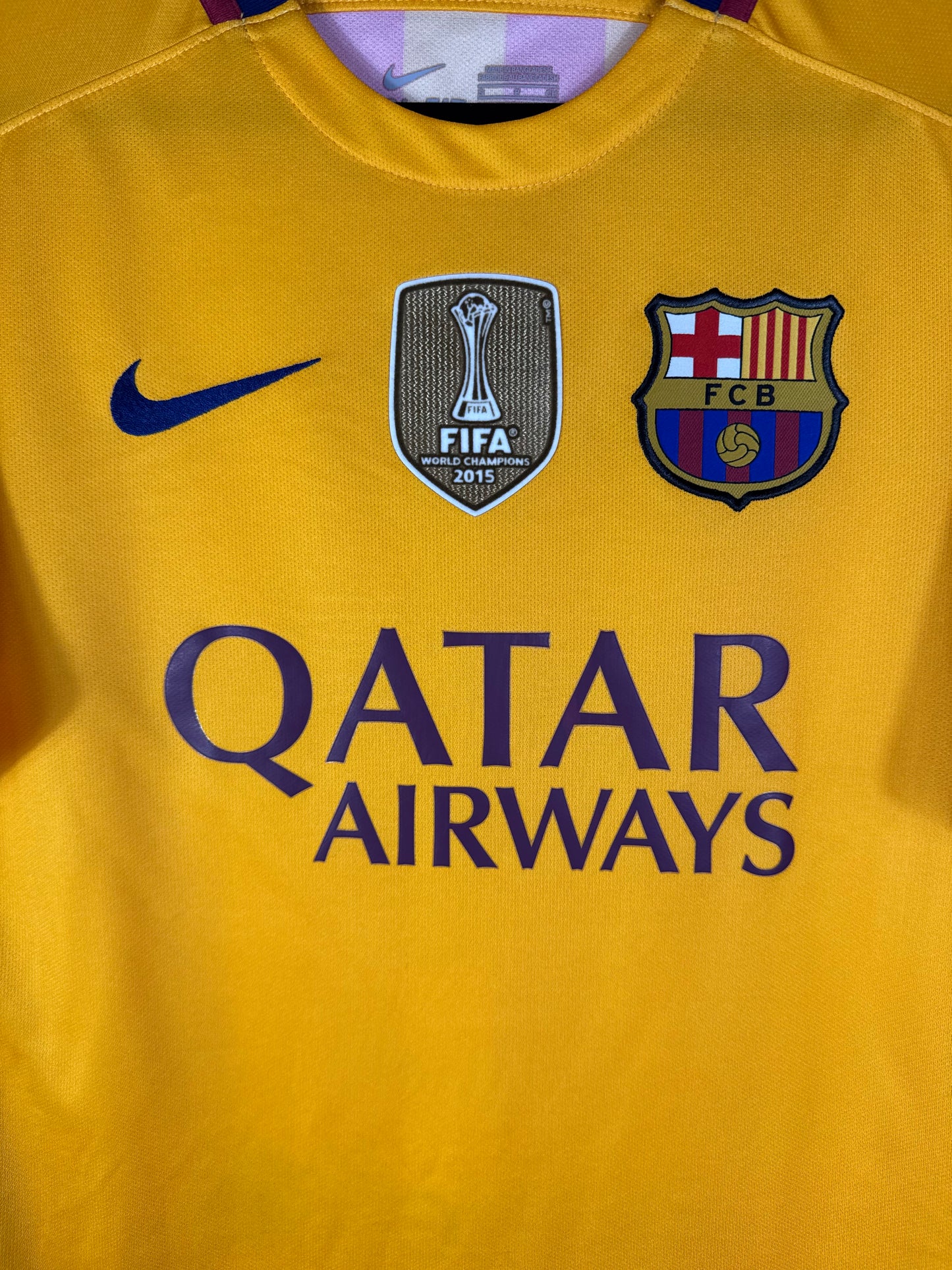 Gros plan sur l'écusson du FC Barcelone, le badge FIFA World Champions doré et le logo Nike bleu sur fond jaune.