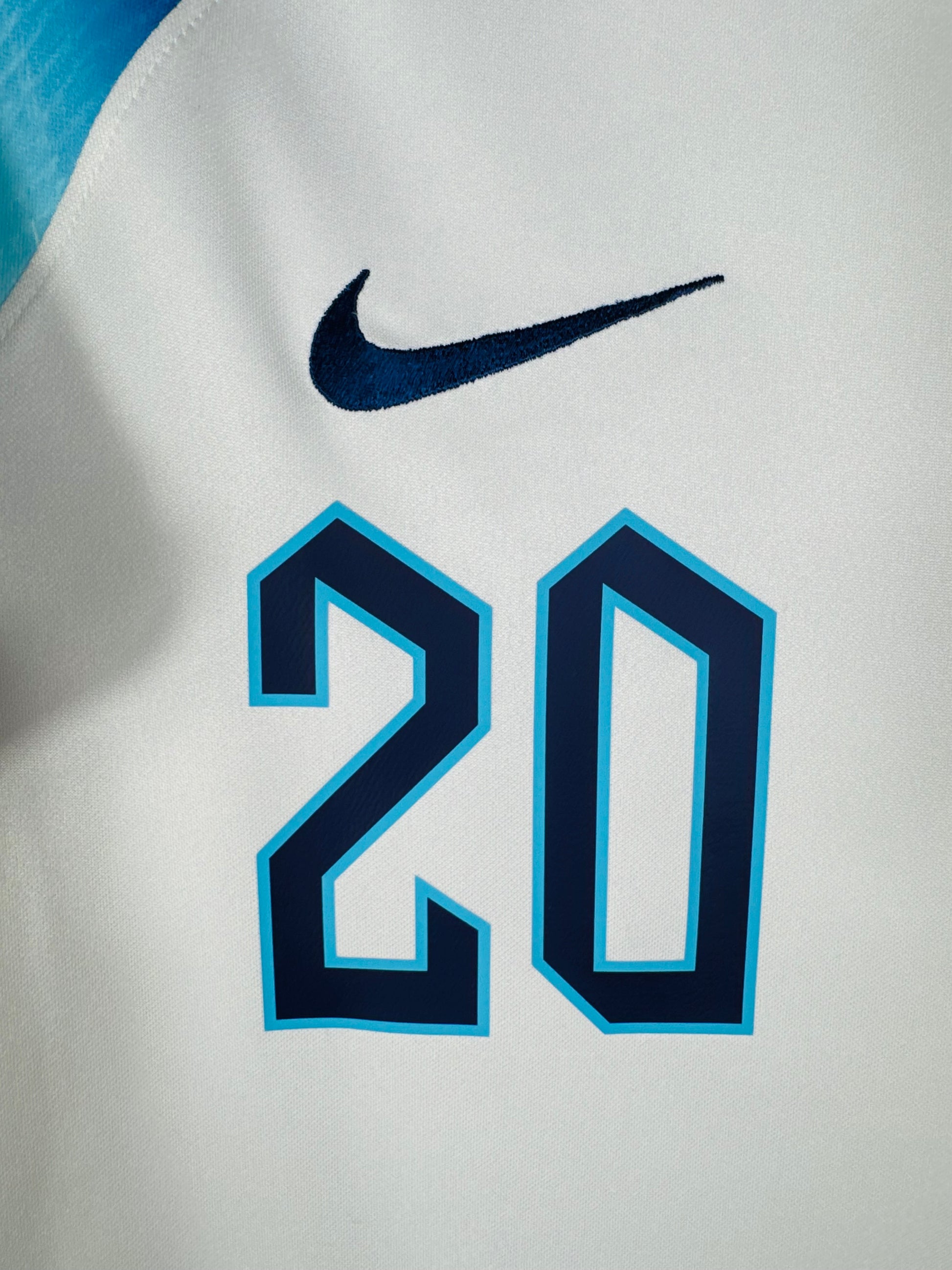 Gros plan sur le blason des Three Lions, le logo Nike bleu marine et le numéro 20 sur le torse.