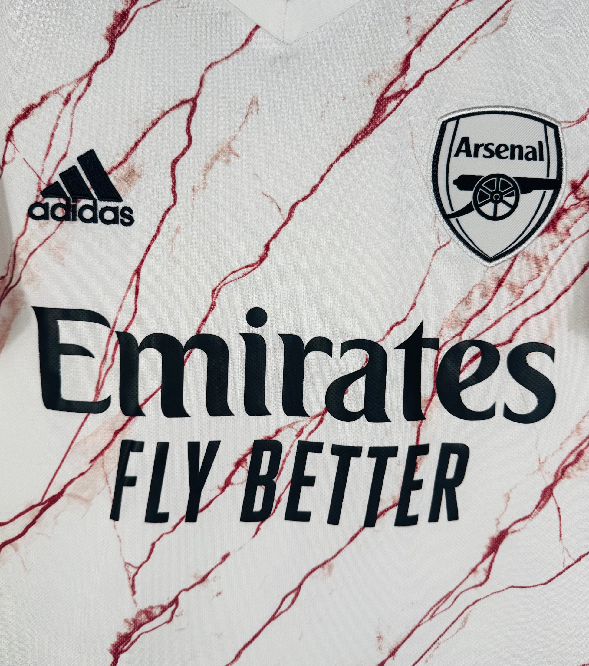 Gros plan sur le logo Adidas et l'écusson Arsenal monochromes noirs sur fond marbré rouge.