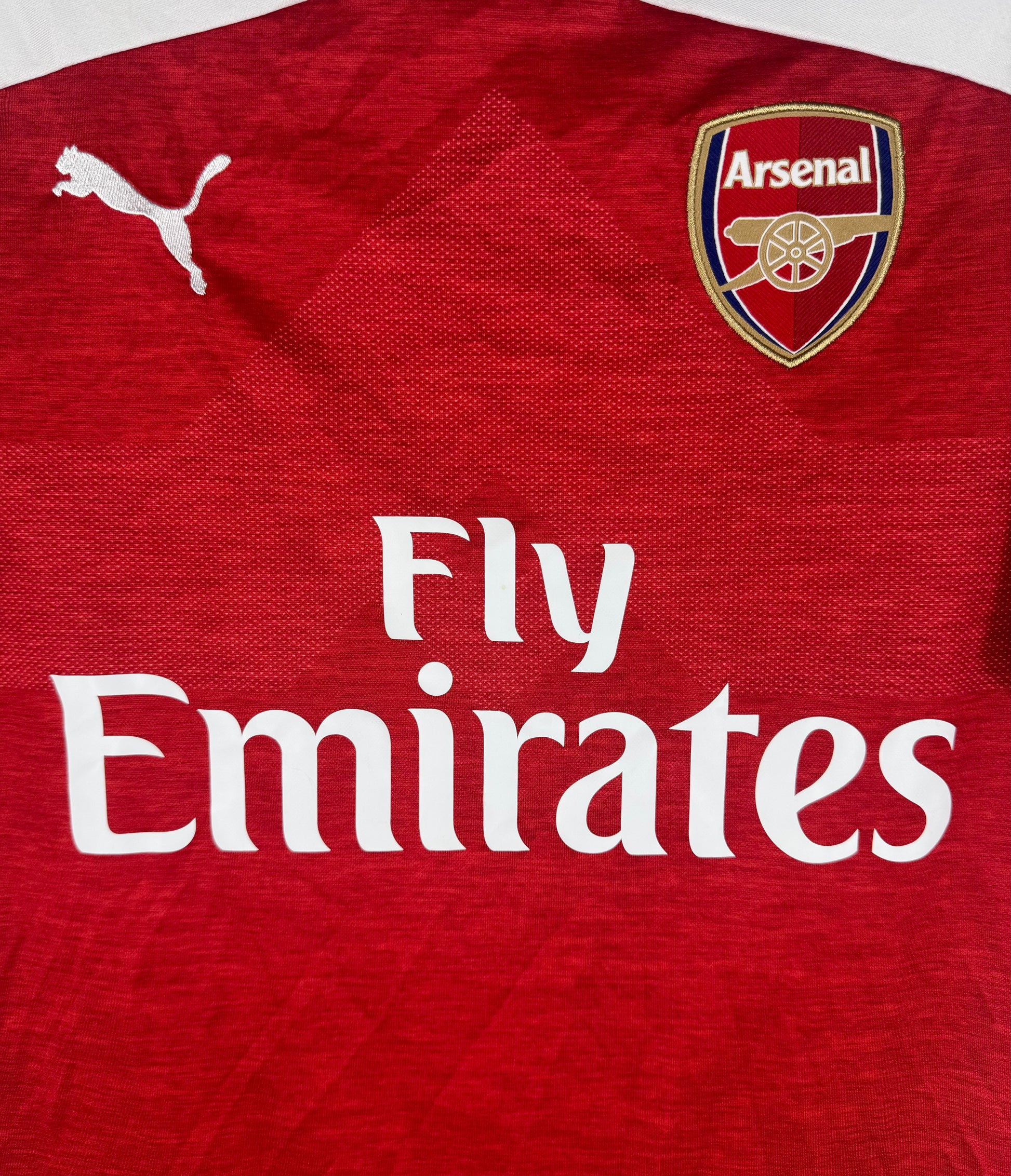 Gros plan sur l'écusson des Gunners et le sponsor Fly Emirates sur fond rouge chiné.