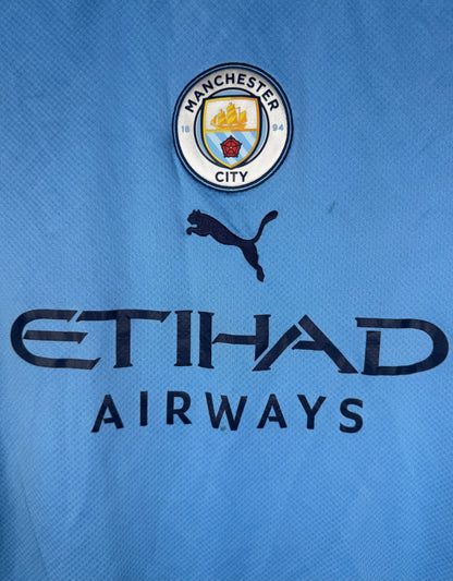 Gros plan sur l'écusson Manchester City centré et le logo Puma sur maillot bleu ciel.