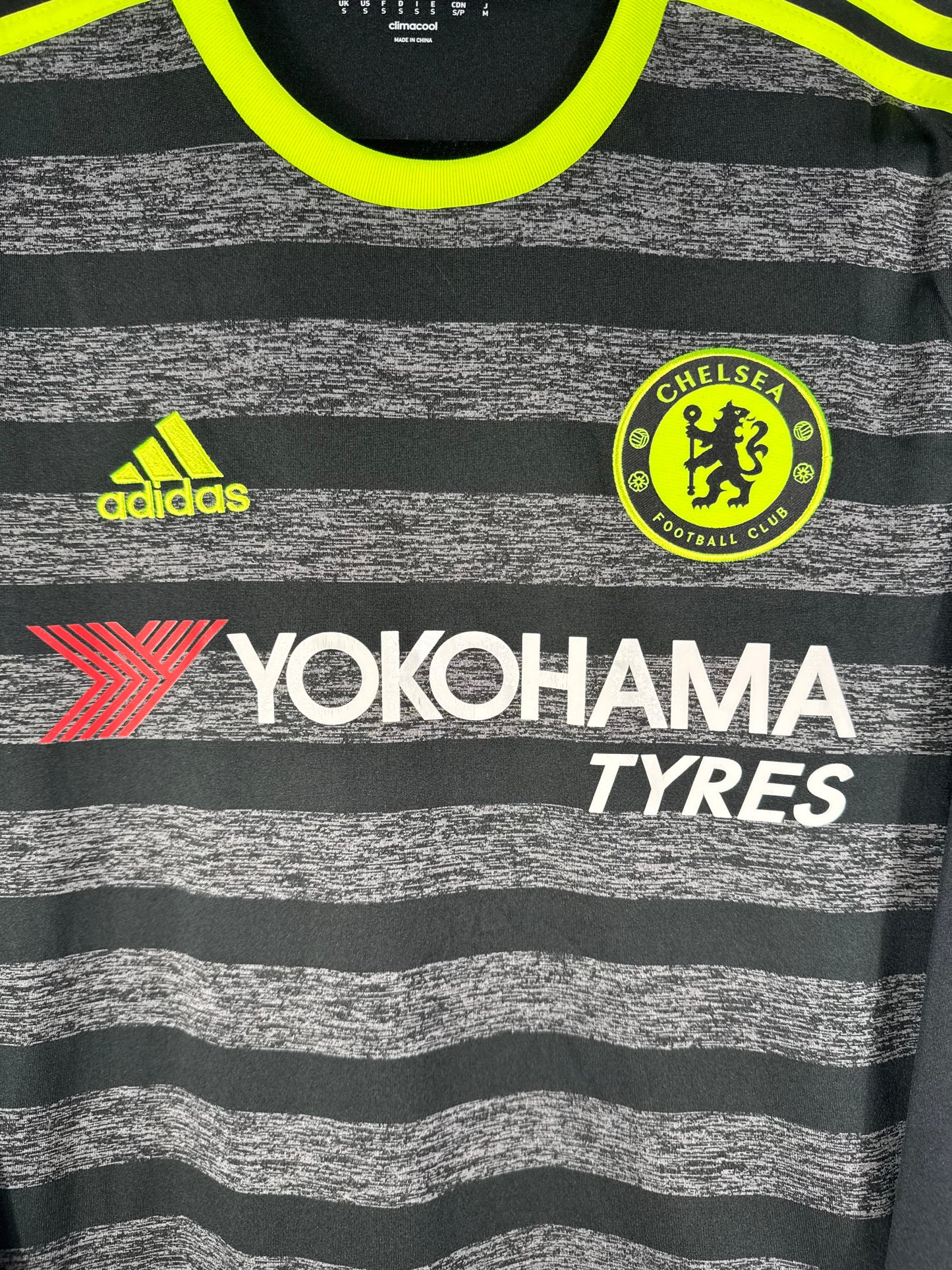 Gros plan sur l'écusson de Chelsea monochrome et le logo Adidas jaune fluo sur fond chiné.