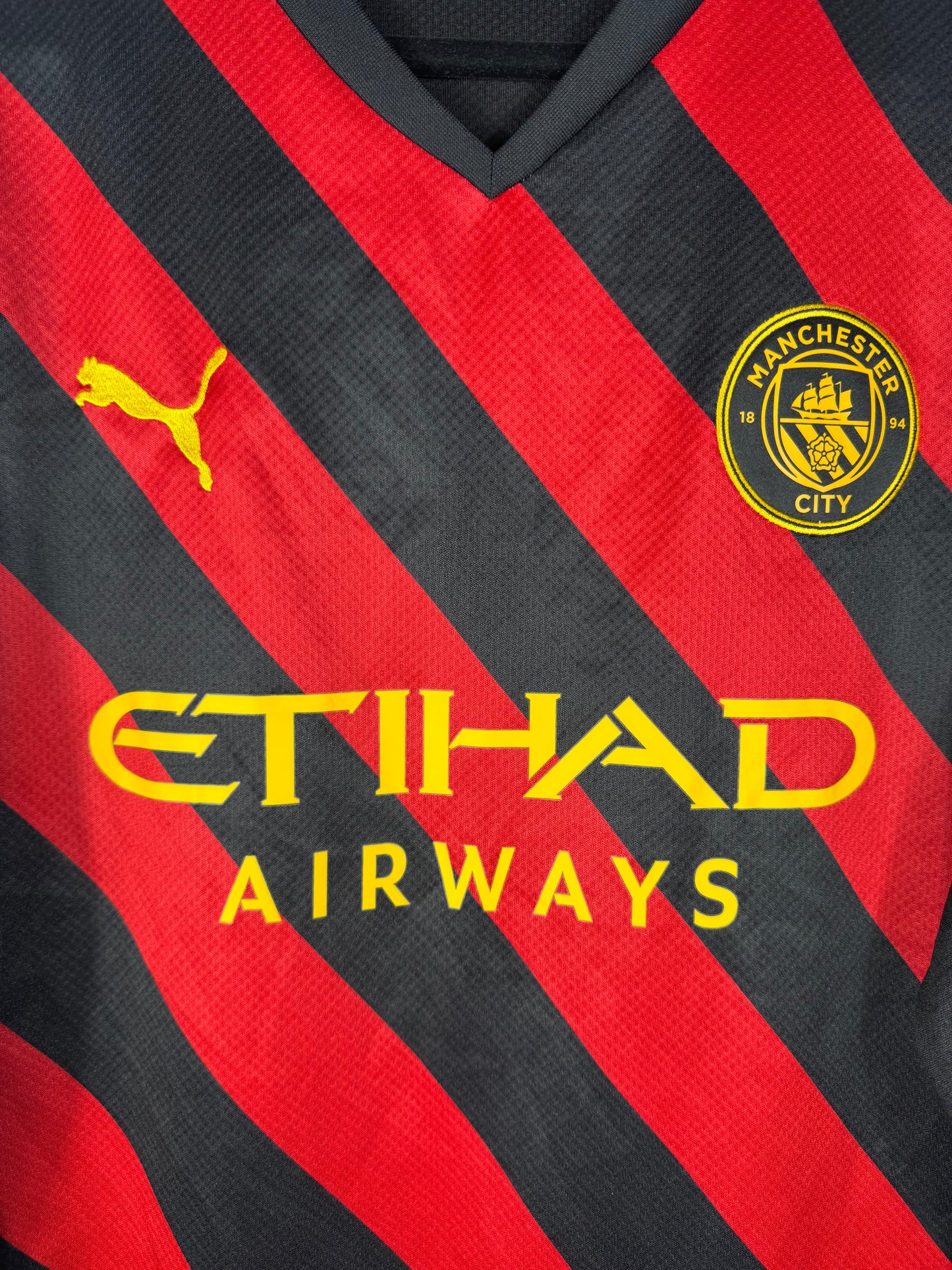 Gros plan sur l'écusson de Manchester City et le logo Puma jaune sur le maillot extérieur à bandes rouges et noires.