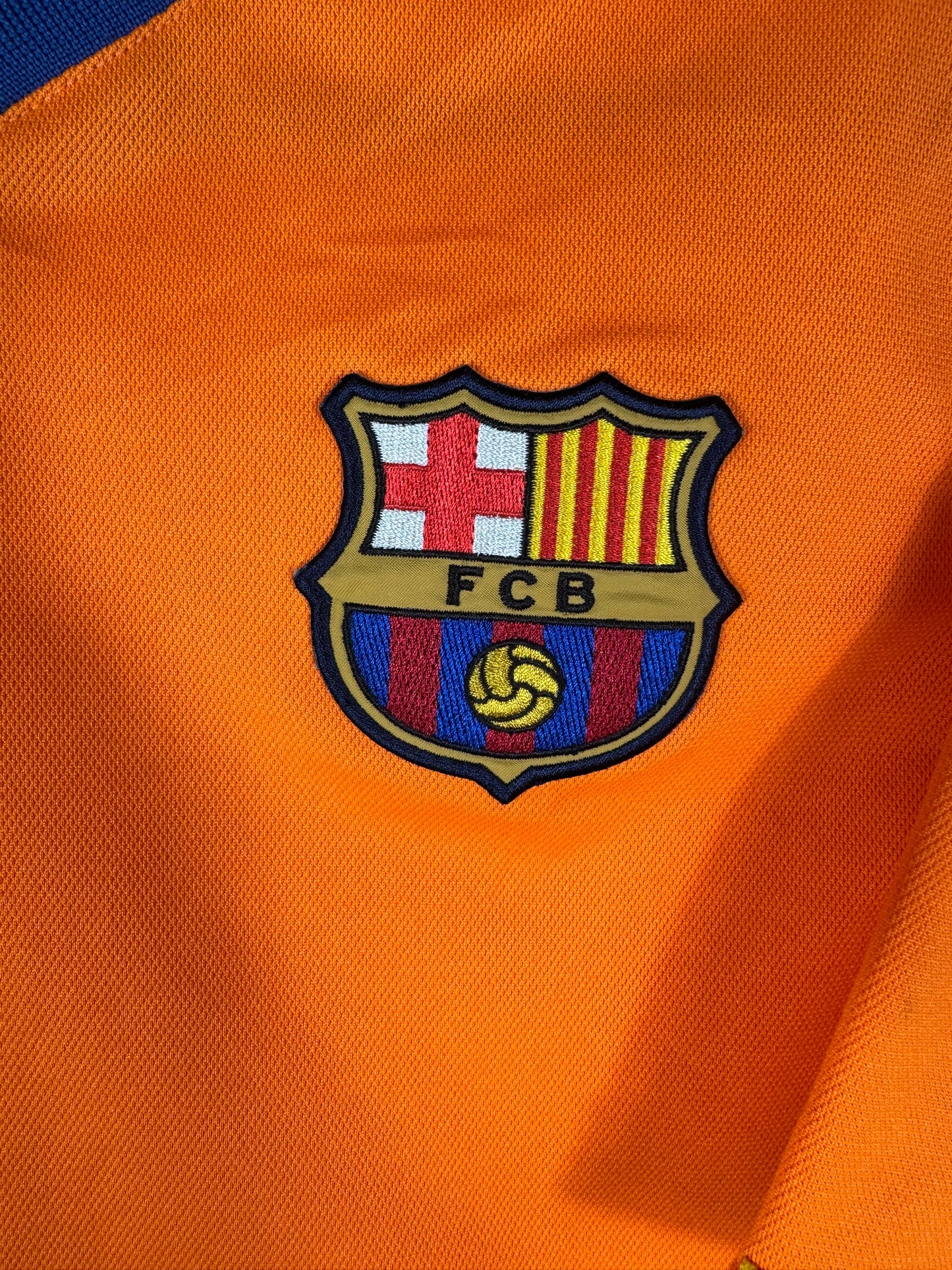 Gros plan sur l'écusson brodé du FC Barcelone sur le torse du maillot extérieur orange.