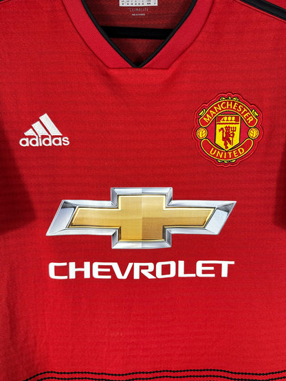 Gros plan sur l'écusson de Manchester United, le logo Adidas blanc et le sponsor Chevrolet sur fond rouge texturé.