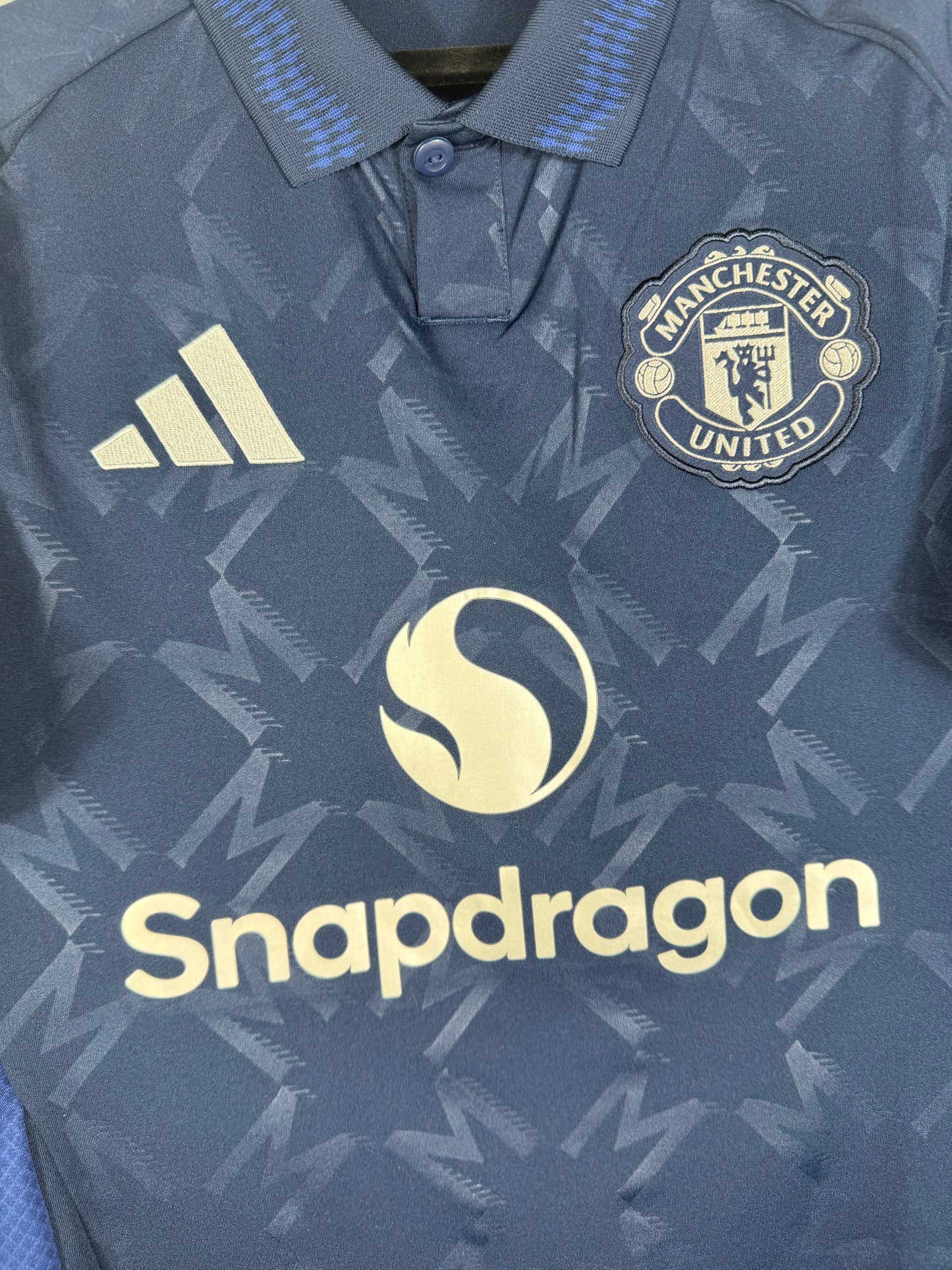 Gros plan sur l'écusson monochrome argenté de Manchester United, le logo Adidas et le sponsor Snapdragon sur le tissu texturé.