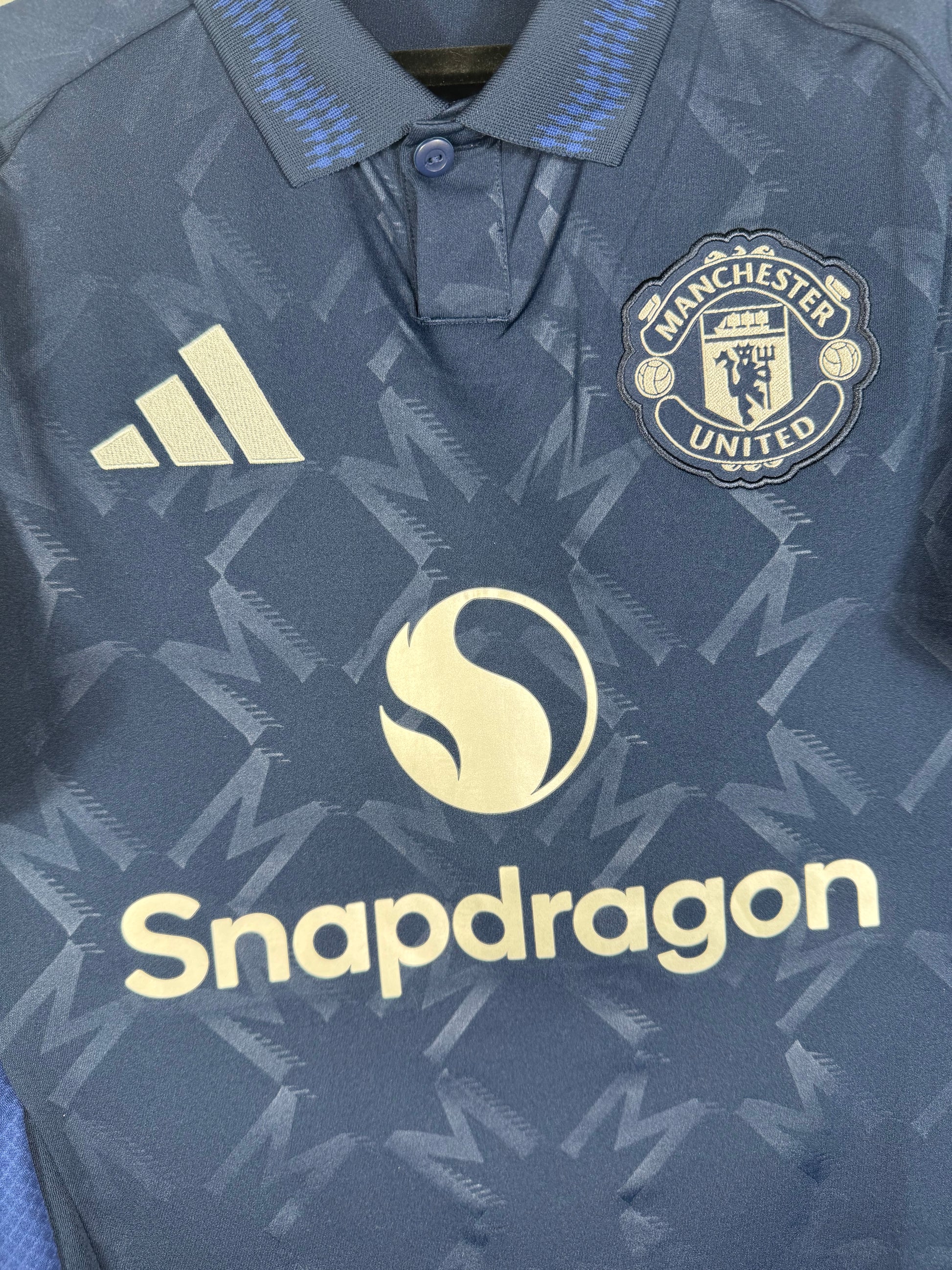 Gros plan sur l'écusson monochrome argenté de Manchester United, le logo Adidas et le sponsor Snapdragon sur le tissu texturé.