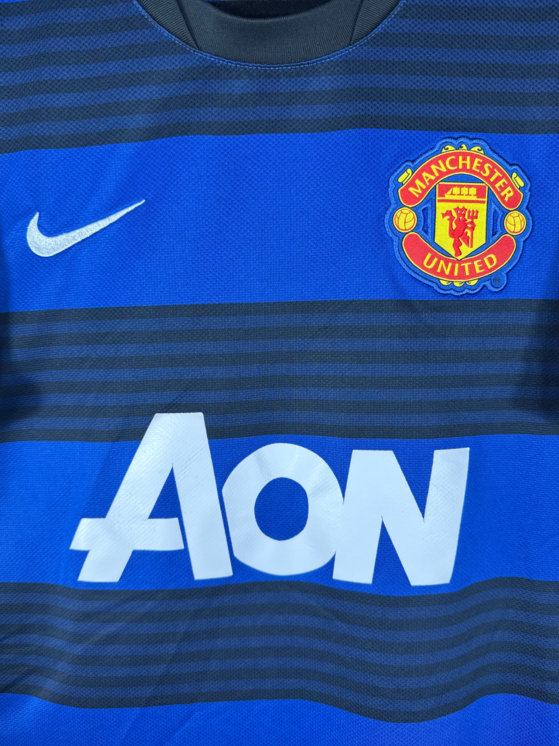 Gros plan sur l'écusson de Manchester United, le logo Nike blanc et le sponsor AON sur les rayures.