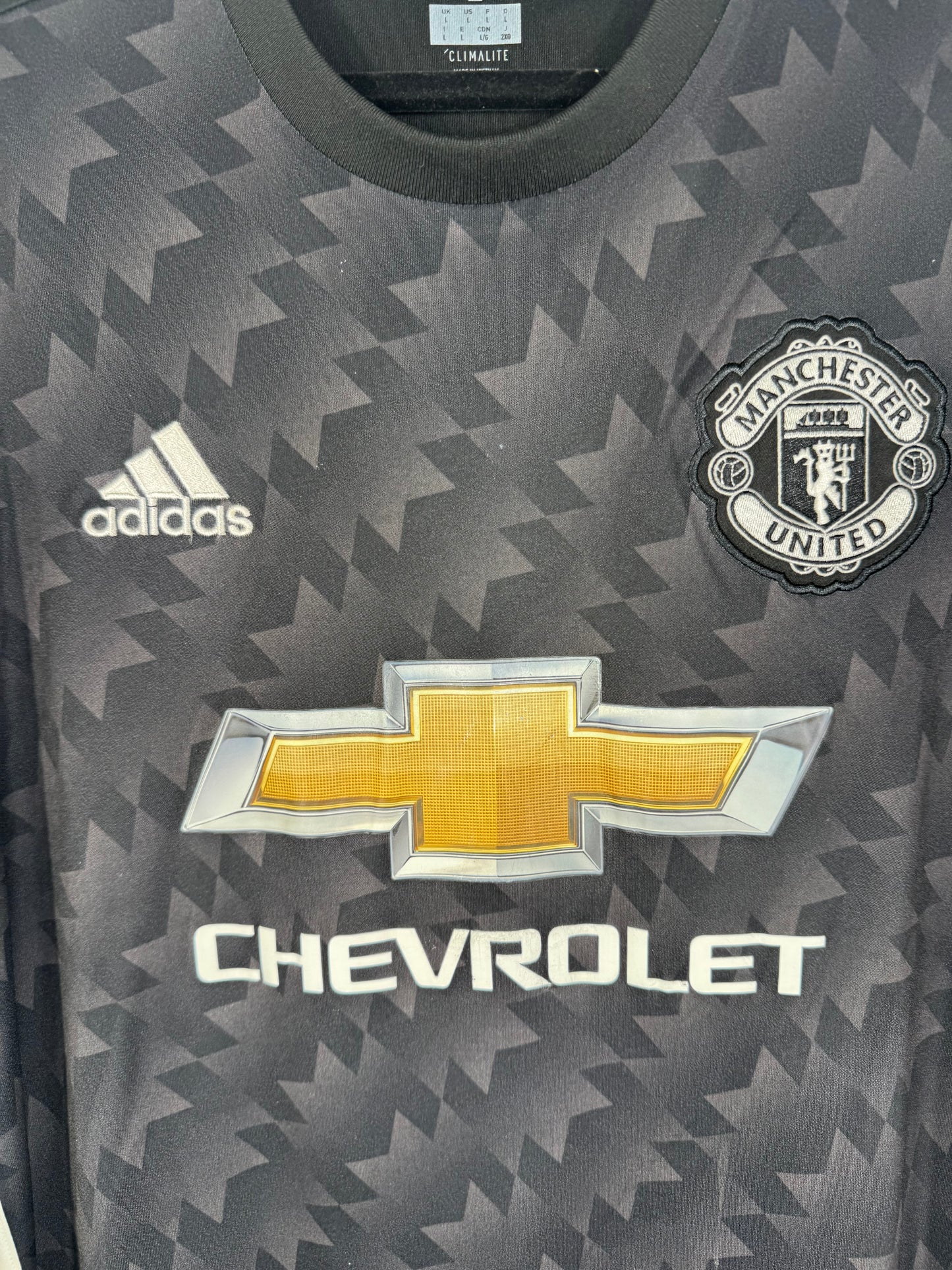 Gros plan sur l'écusson de Manchester United et le logo Adidas brodés en blanc sur fond noir.