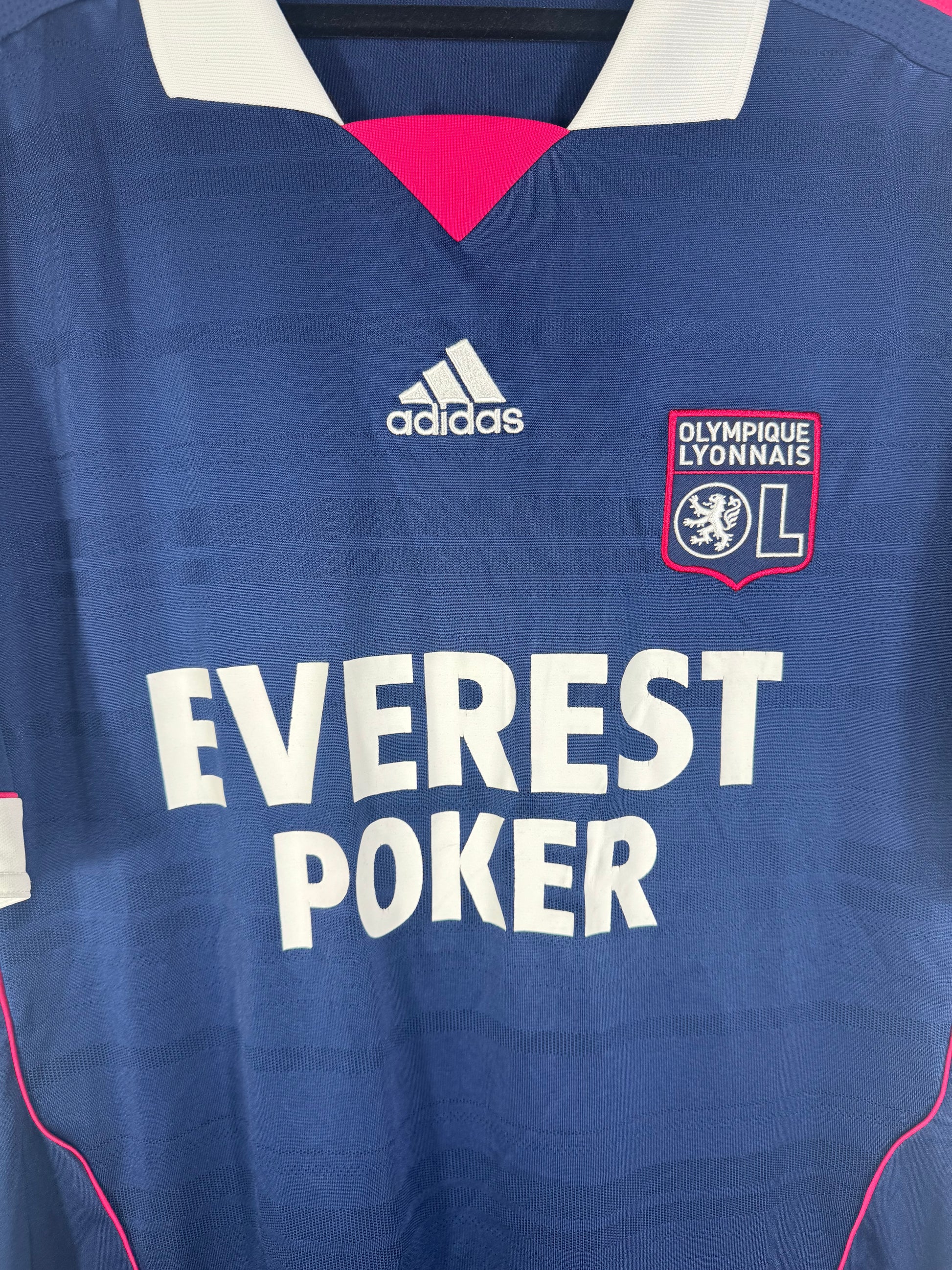 Gros plan sur l'écusson de l'OL, le logo Adidas et le sponsor Everest Poker sur le tissu texturé bleu.