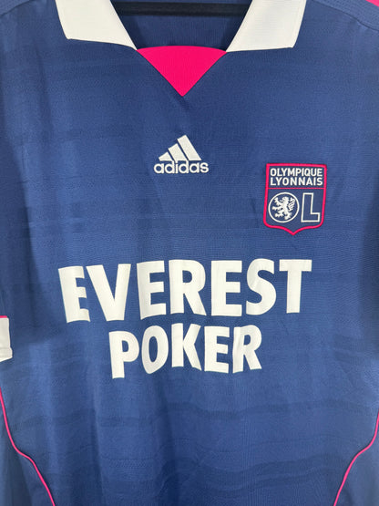 Gros plan sur l'écusson de l'OL, le logo Adidas et le sponsor Everest Poker sur le tissu texturé bleu.