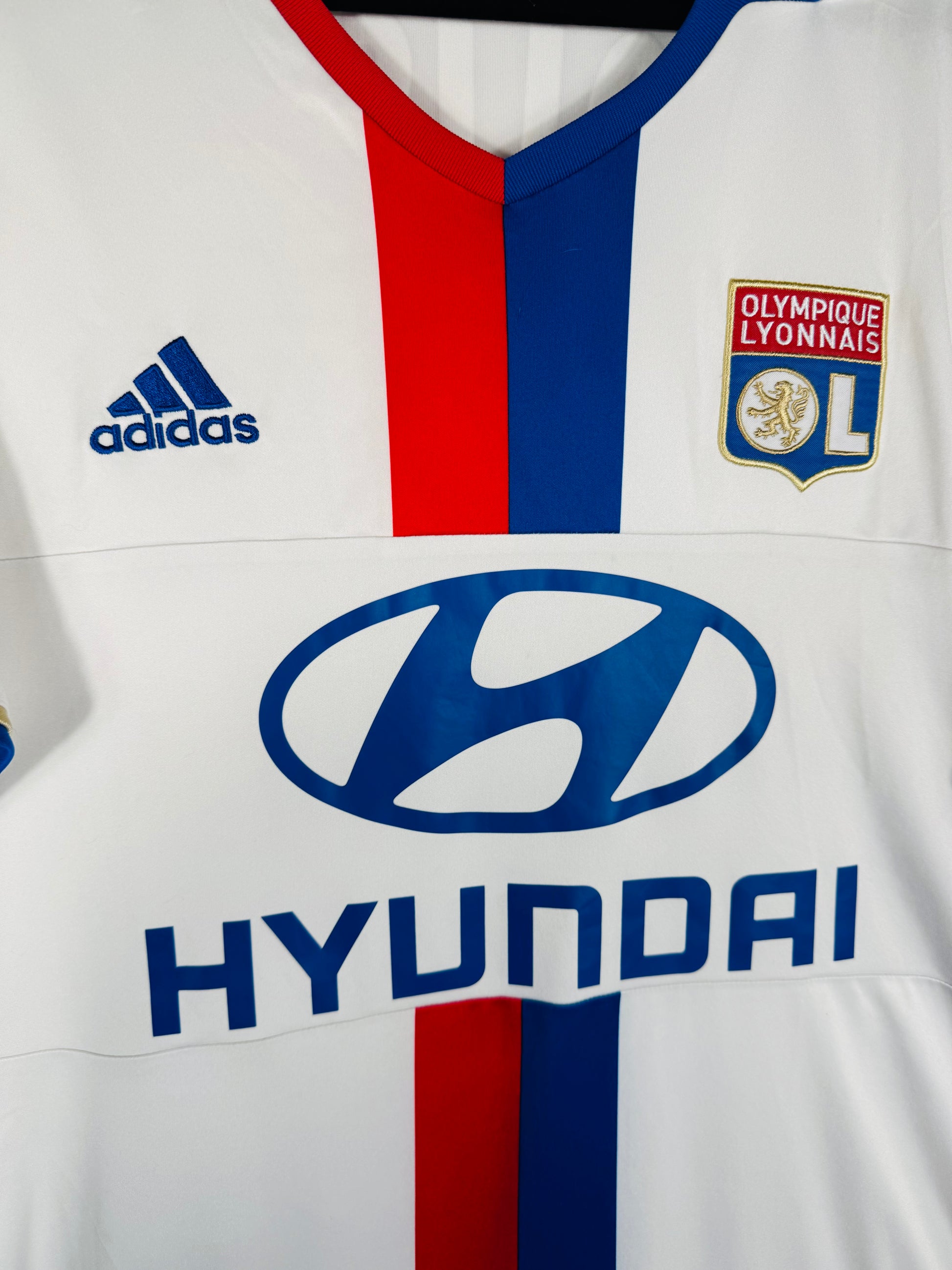 Gros plan sur l'écusson de l'OL et le logo Adidas brodé en fil doré.
