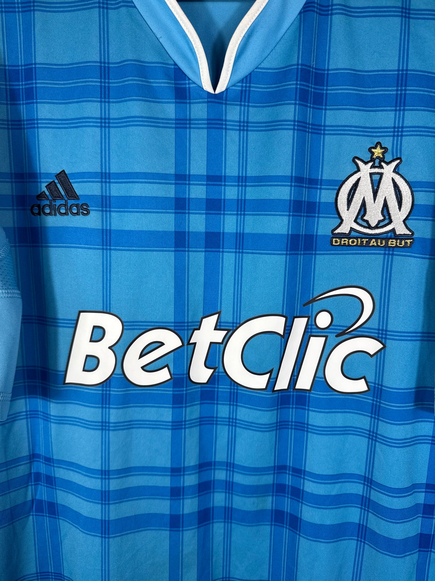 Gros plan sur l'écusson de l'OM et le logo Adidas sur le maillot extérieur à motifs tartan.