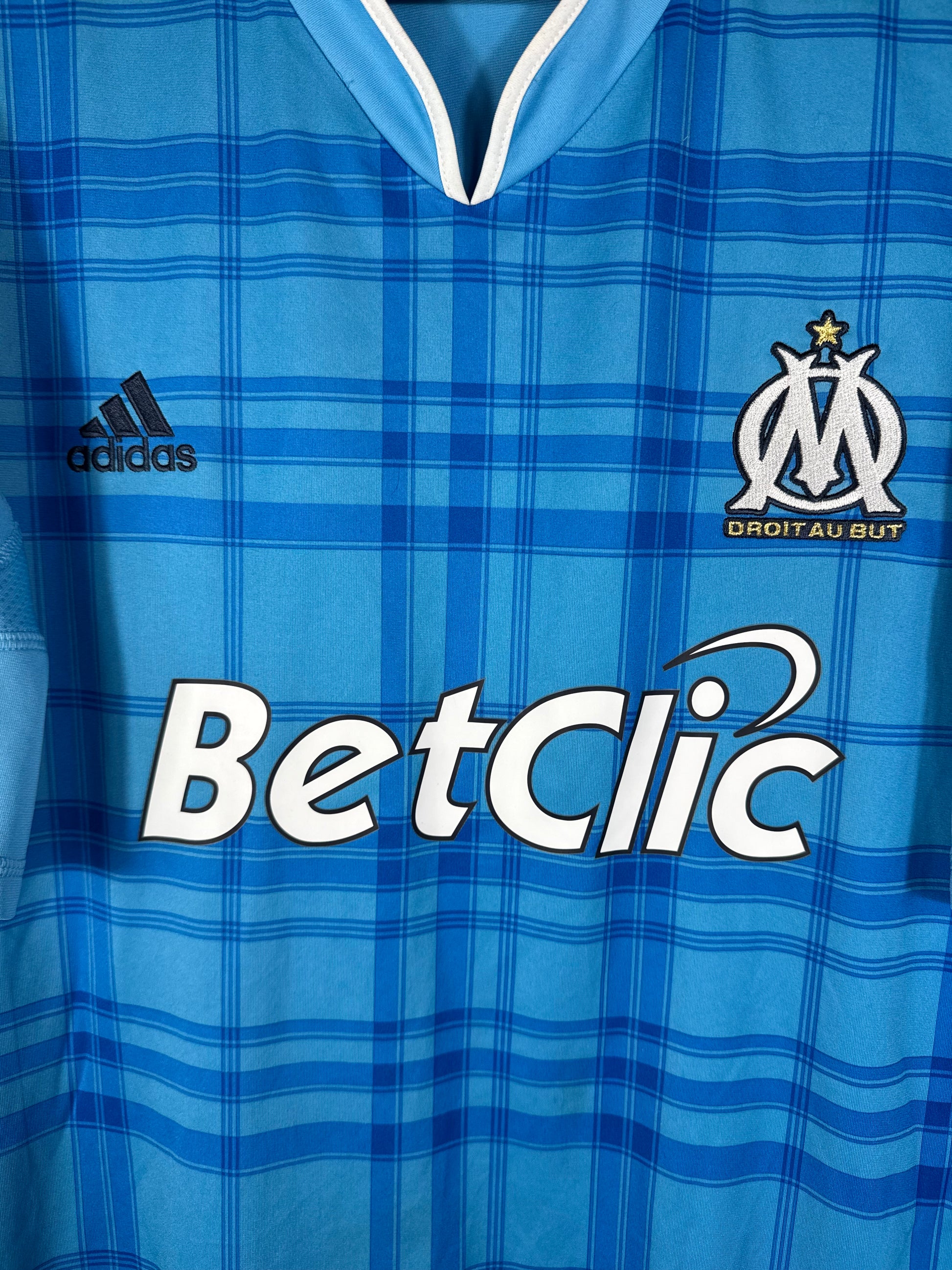 Gros plan sur l'écusson de l'OM et le logo Adidas sur le maillot extérieur à motifs tartan.