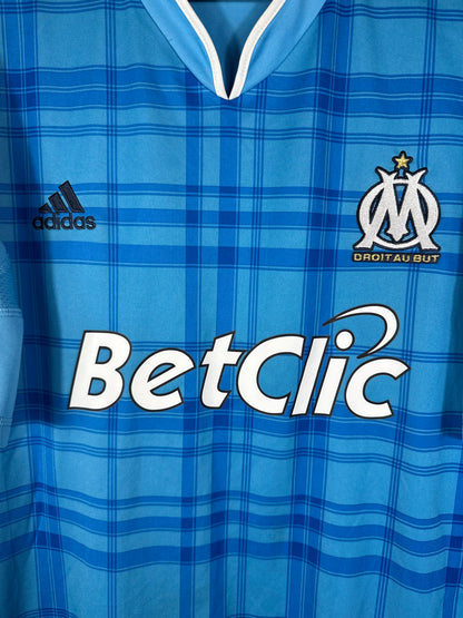 Gros plan sur l'écusson de l'OM et le logo Adidas sur le maillot extérieur à motifs tartan.