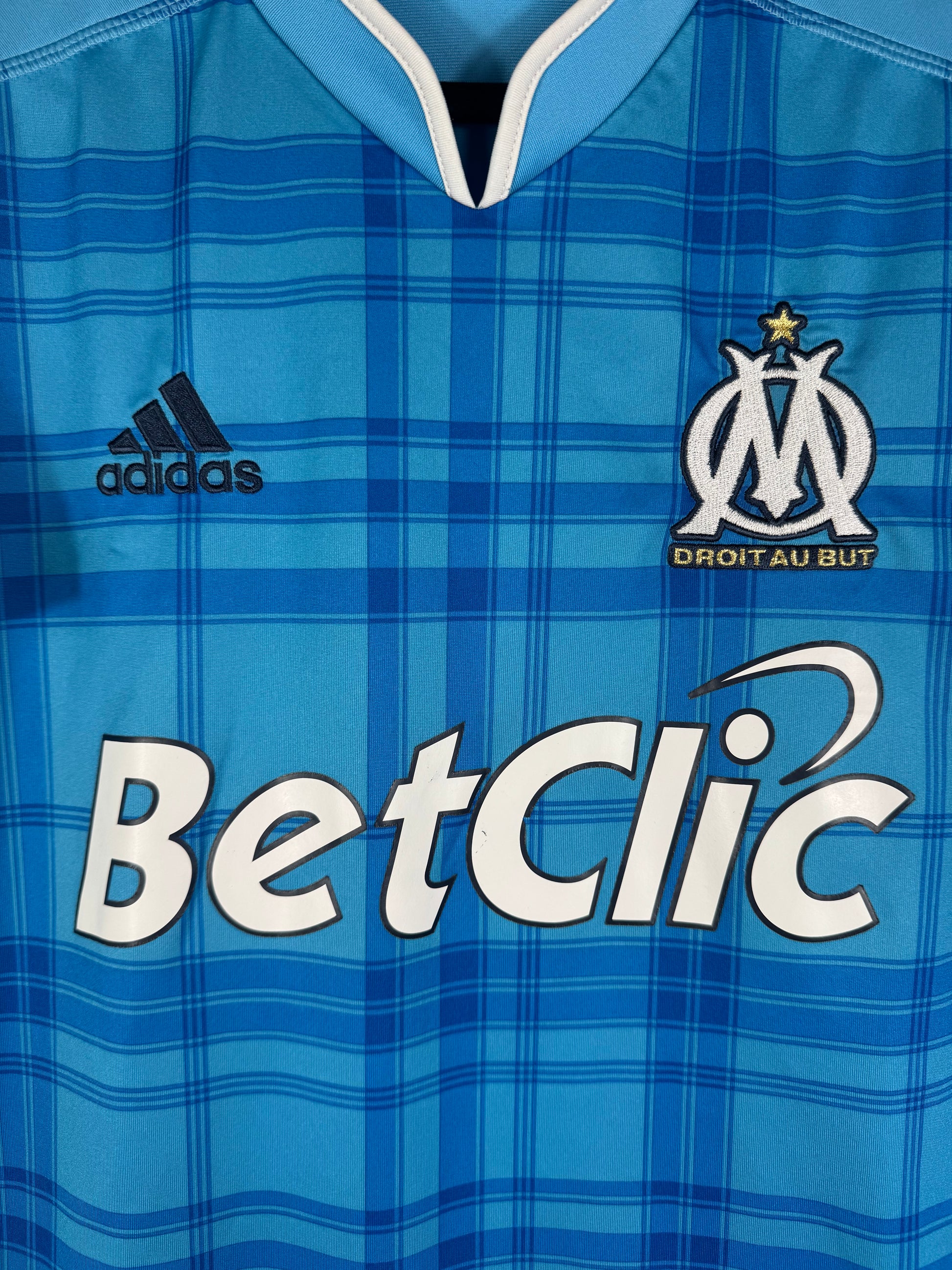Gros plan sur l'écusson de l'OM, le logo Adidas bleu marine et le sponsor BetClic sur le tissu à carreaux.