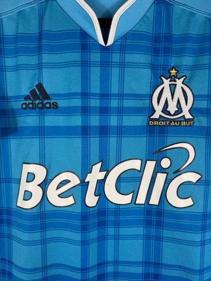 Gros plan sur l'écusson de l'OM, le logo Adidas bleu marine et le sponsor BetClic sur le tissu à carreaux.