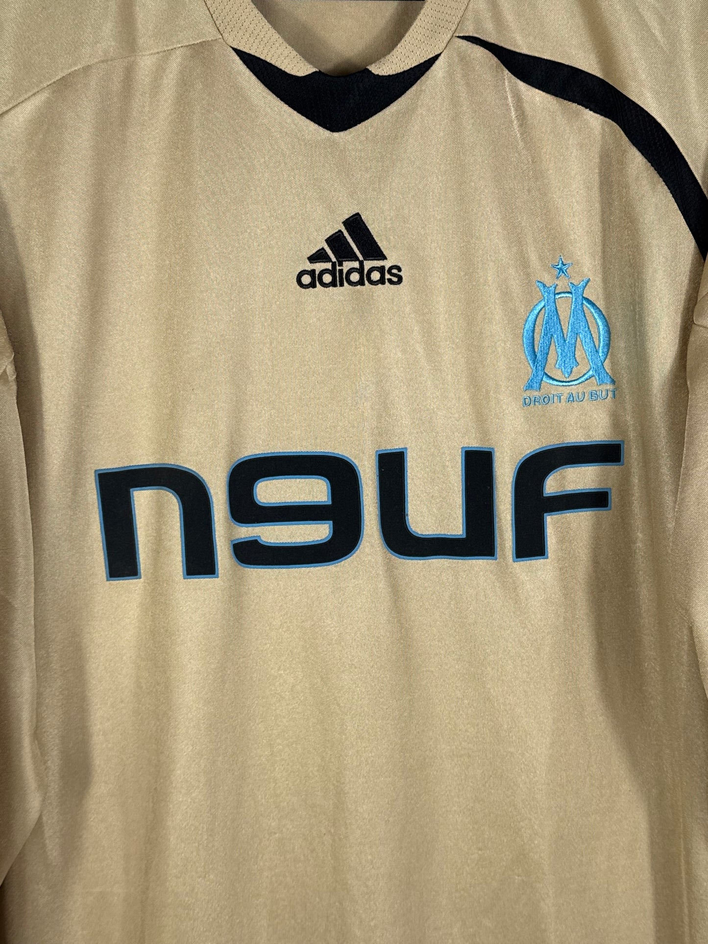 Gros plan sur l'écusson de l'OM, le logo Adidas et le sponsor n9uf sur le maillot doré.