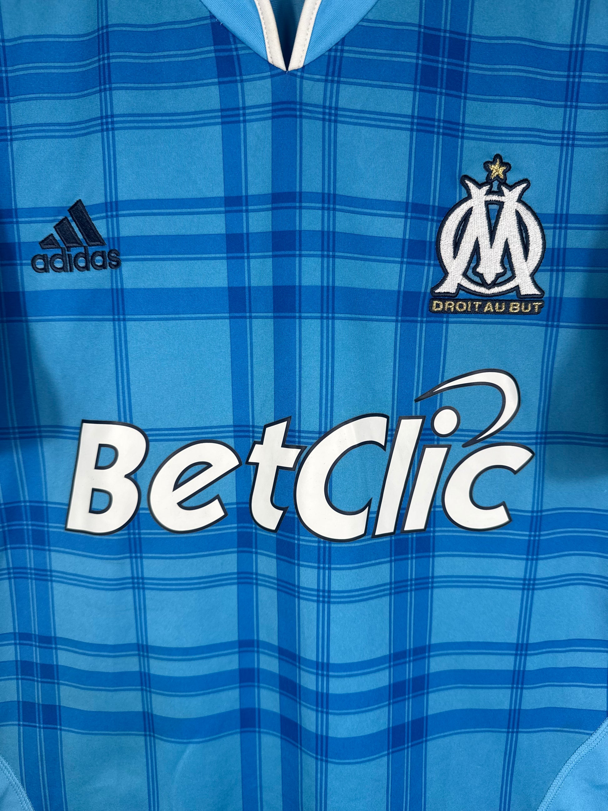 Gros plan sur l'écusson Olympique de Marseille doré et le motif bleu quadrillé.