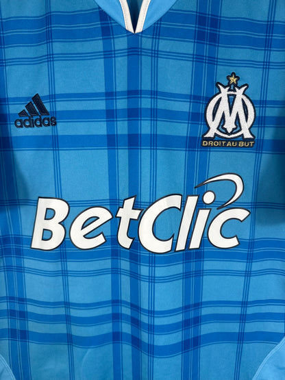 Gros plan sur l'écusson Olympique de Marseille doré et le motif bleu quadrillé.