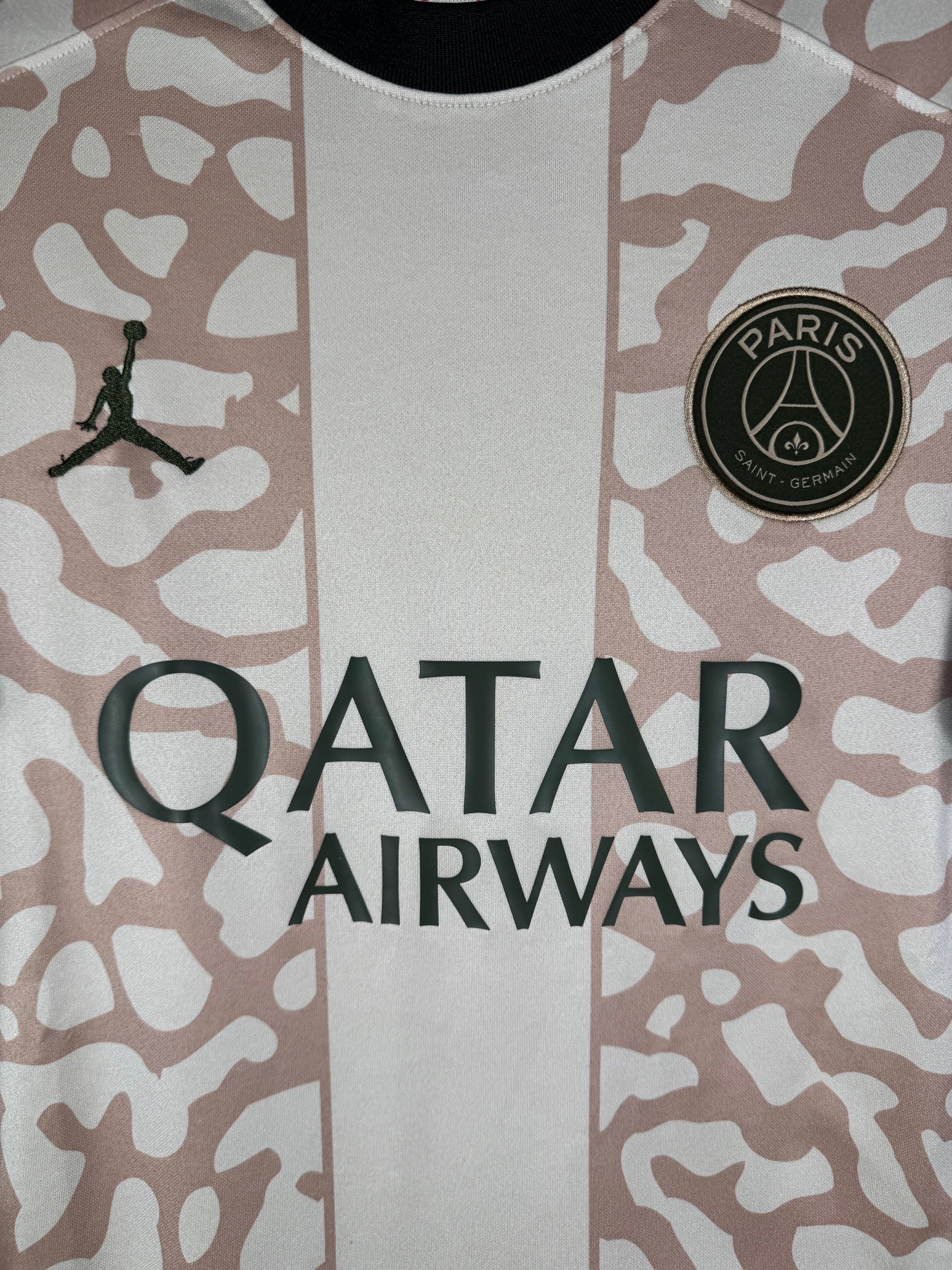 Gros plan sur l'écusson du Paris Saint-Germain et le logo Jumpman bleu marine sur le tissu beige.