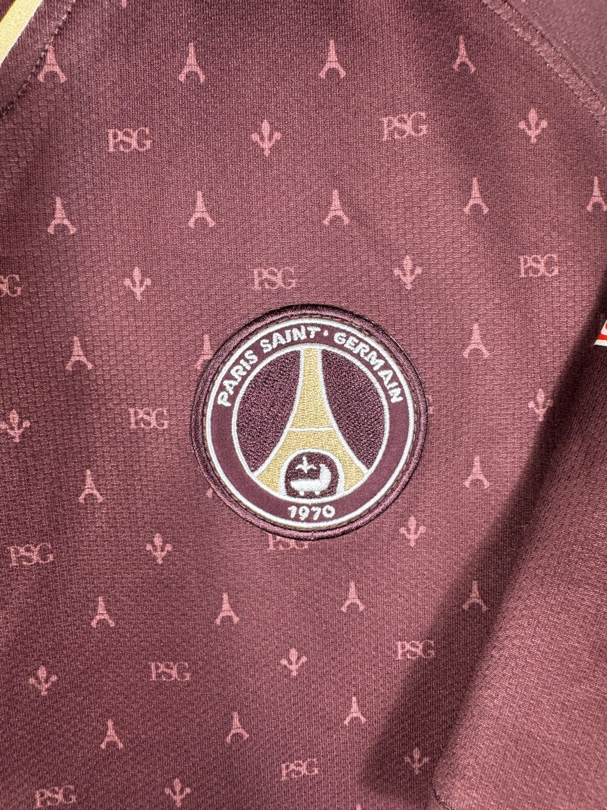 Gros plan sur l'écusson vintage du Paris Saint-Germain fondu dans le motif monogramme doré.