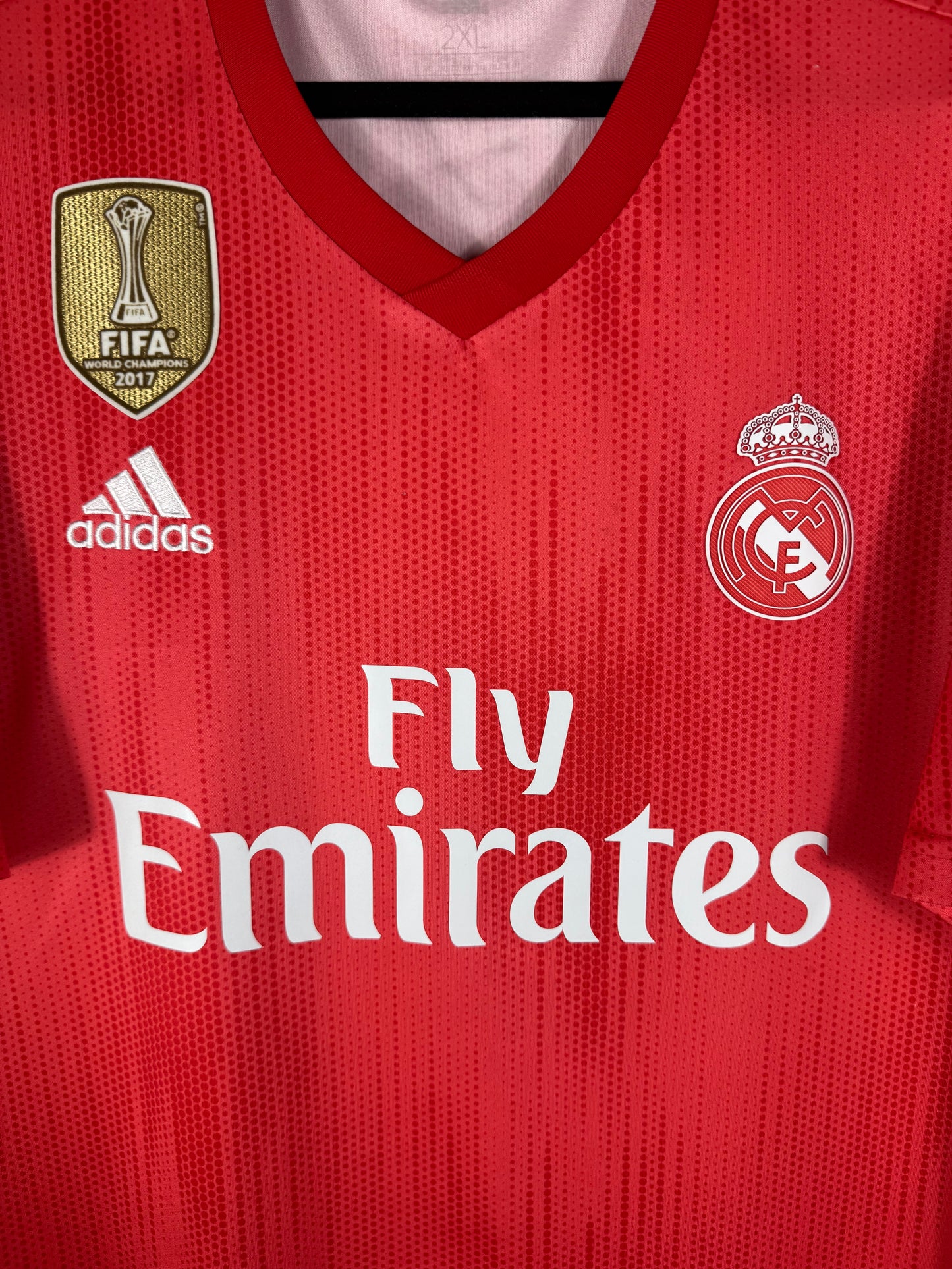 Gros plan sur l'écusson blanc du Real Madrid, le badge FIFA doré et le logo Adidas sur le tissu rouge recyclé.