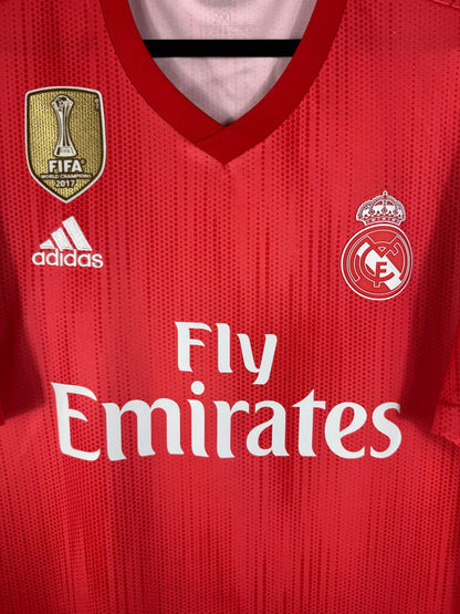 Gros plan sur l'écusson blanc du Real Madrid, le badge FIFA doré et le logo Adidas sur le tissu rouge recyclé.