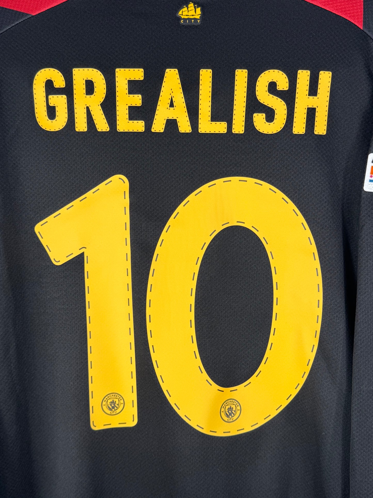 Zoom sur le numéro 10 jaune de Jack Grealish avec typographie officielle Ligue des Champions de Manchester City.
