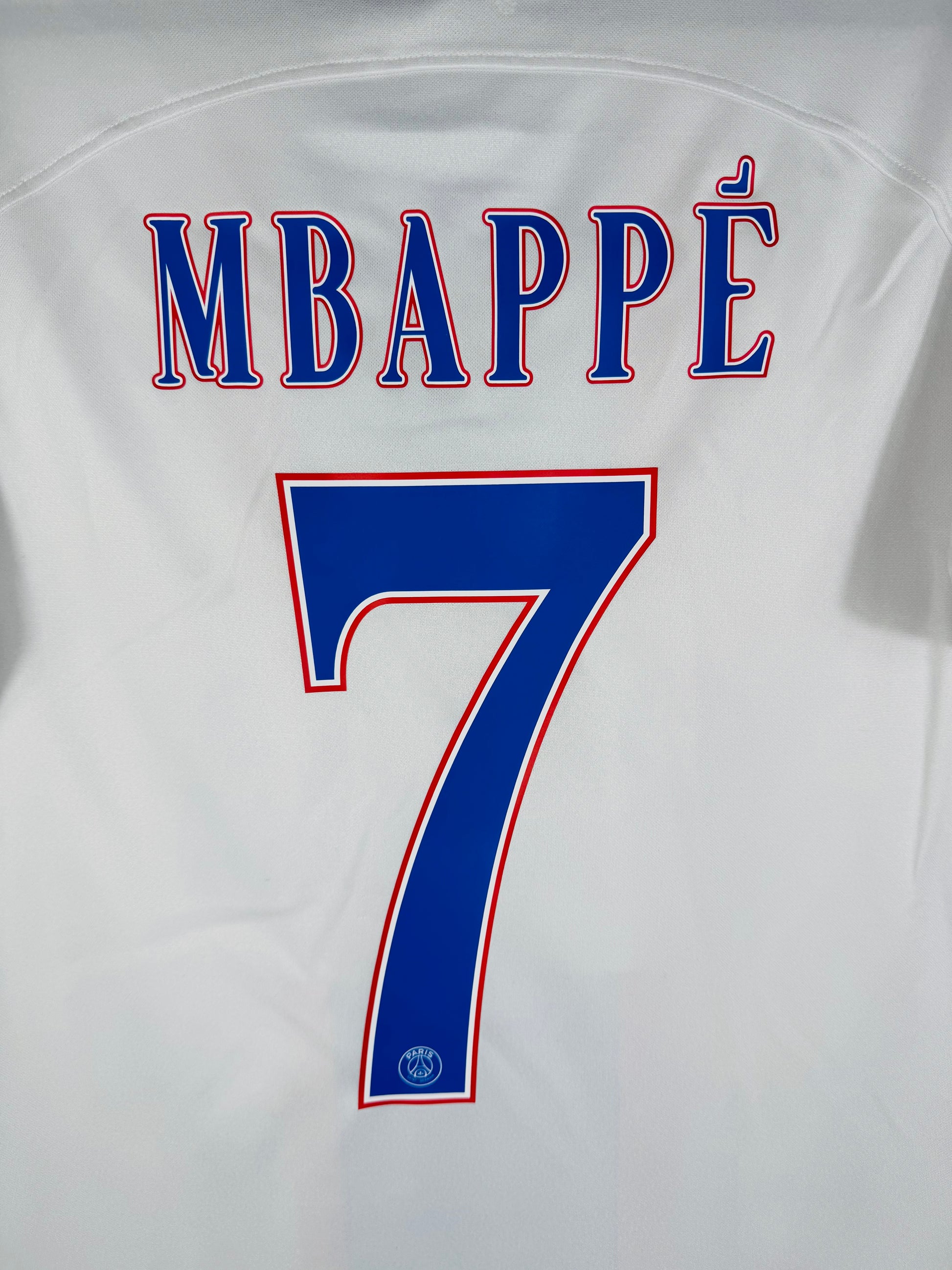 Zoom sur le nom MBAPPÉ et le numéro 7 bleu avec le logo du PSG incrusté au bas du chiffre.