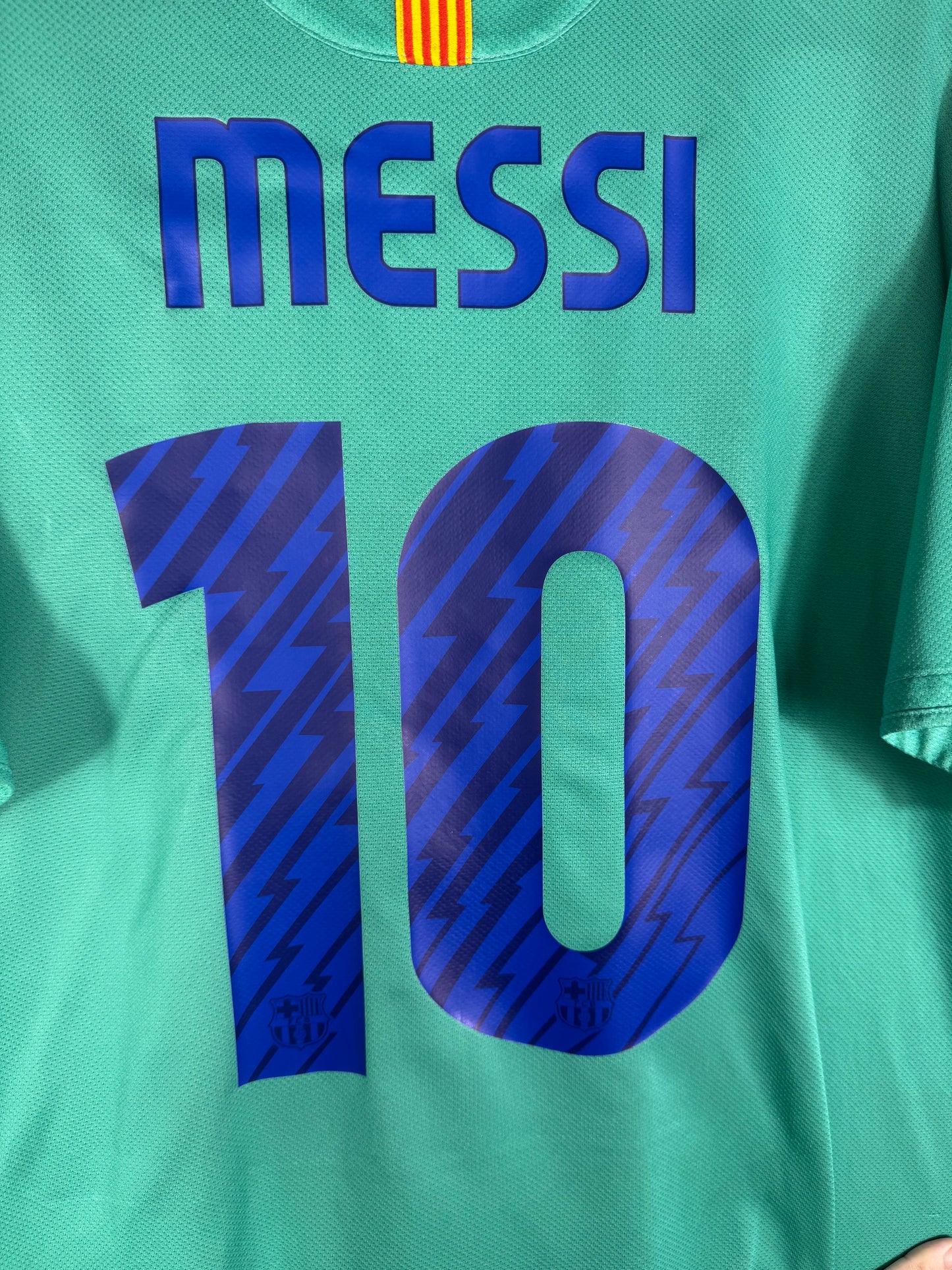 Zoom sur le nom MESSI et le numéro 10 bleu avec motifs à rayures diagonales intégrées, typographie officielle du Barça.