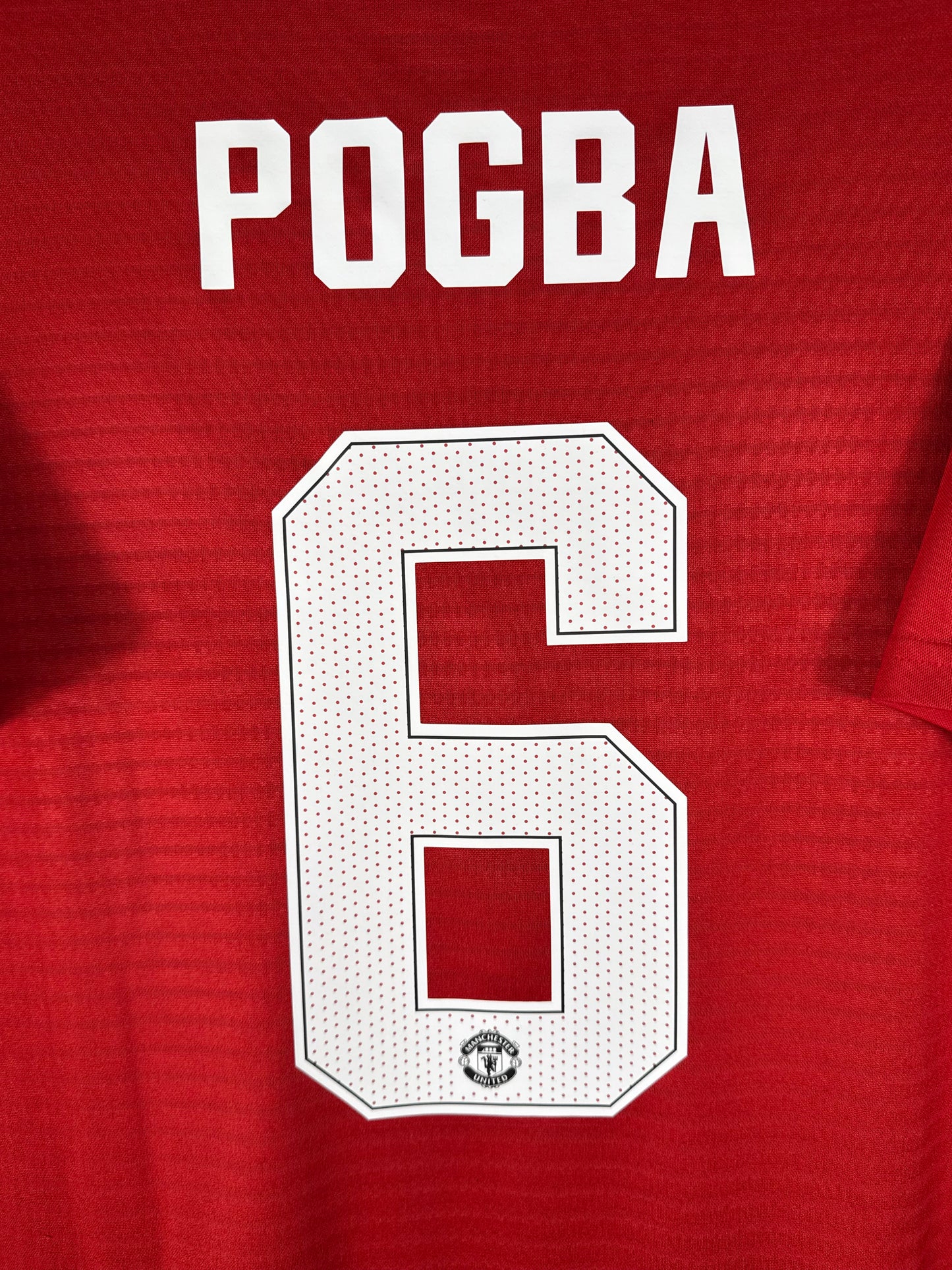 Zoom sur le nom POGBA et le numéro 6 blanc, typographie officielle Manchester United pour les Coupes avec le blason intégré.