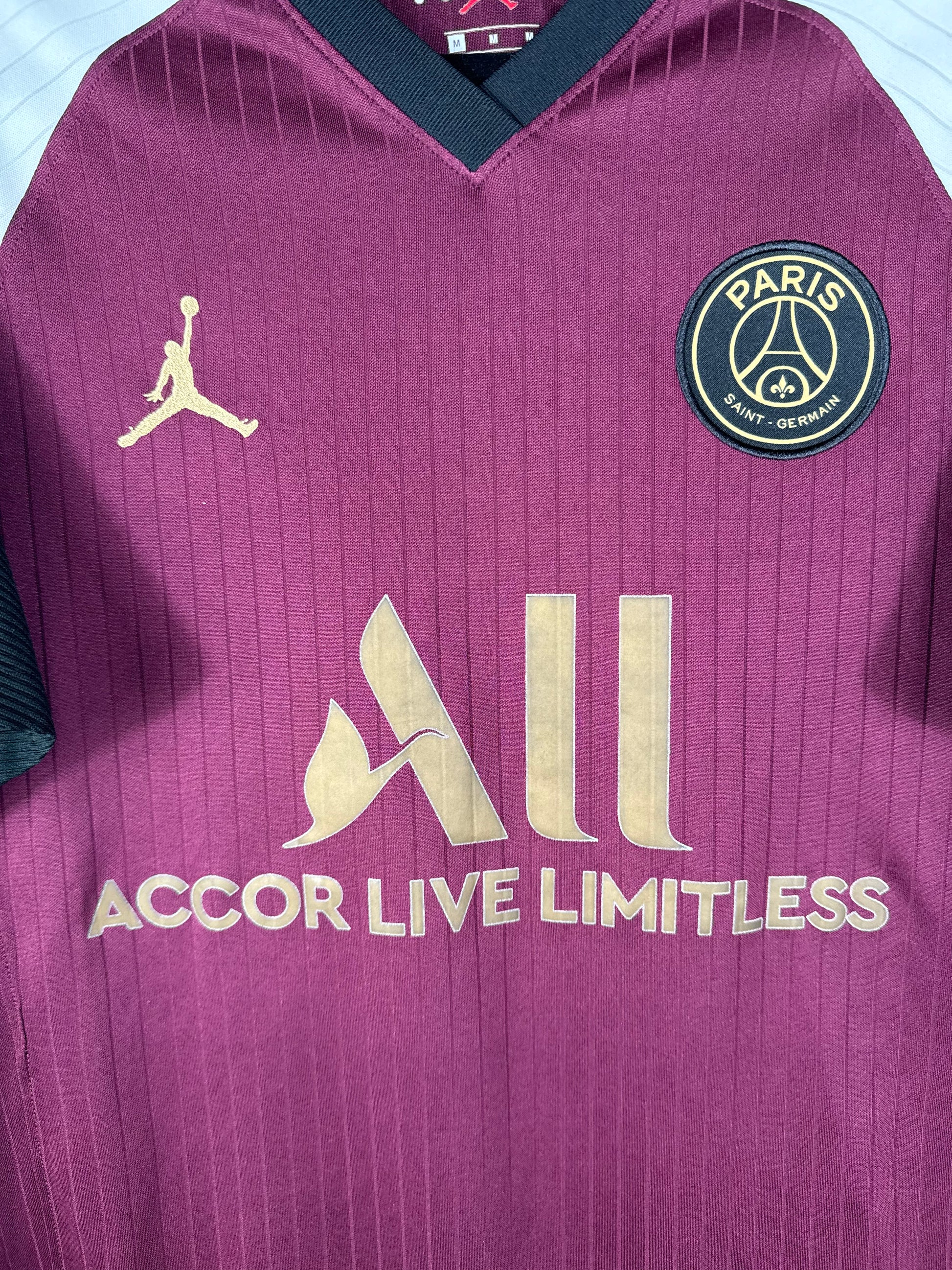 Gros plan sur l'écusson du PSG et le logo Jumpman dorés sur le tissu bordeaux texturé.