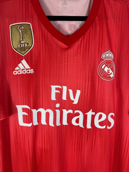 Gros plan sur l'écusson monochrome du Real Madrid, le badge FIFA doré et le logo Adidas blanc.