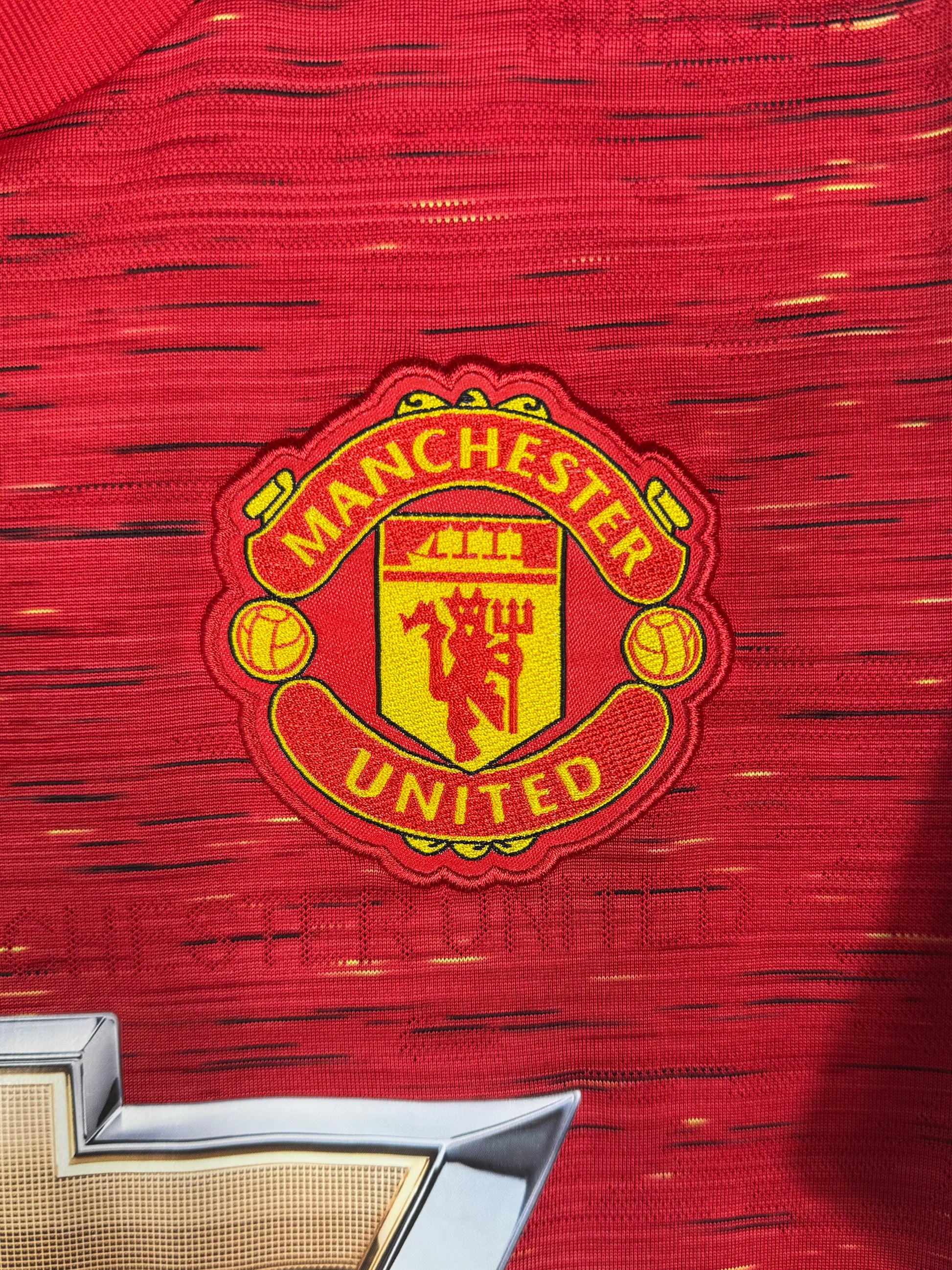 Détail écusson Manchester United brodé sur fond motif tissé rouge et jaune.