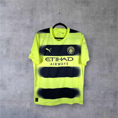 Vue de face du maillot Manchester City Third 2022/23 jaune fluo traversé par des bandes noires horizontales à effet fondu.