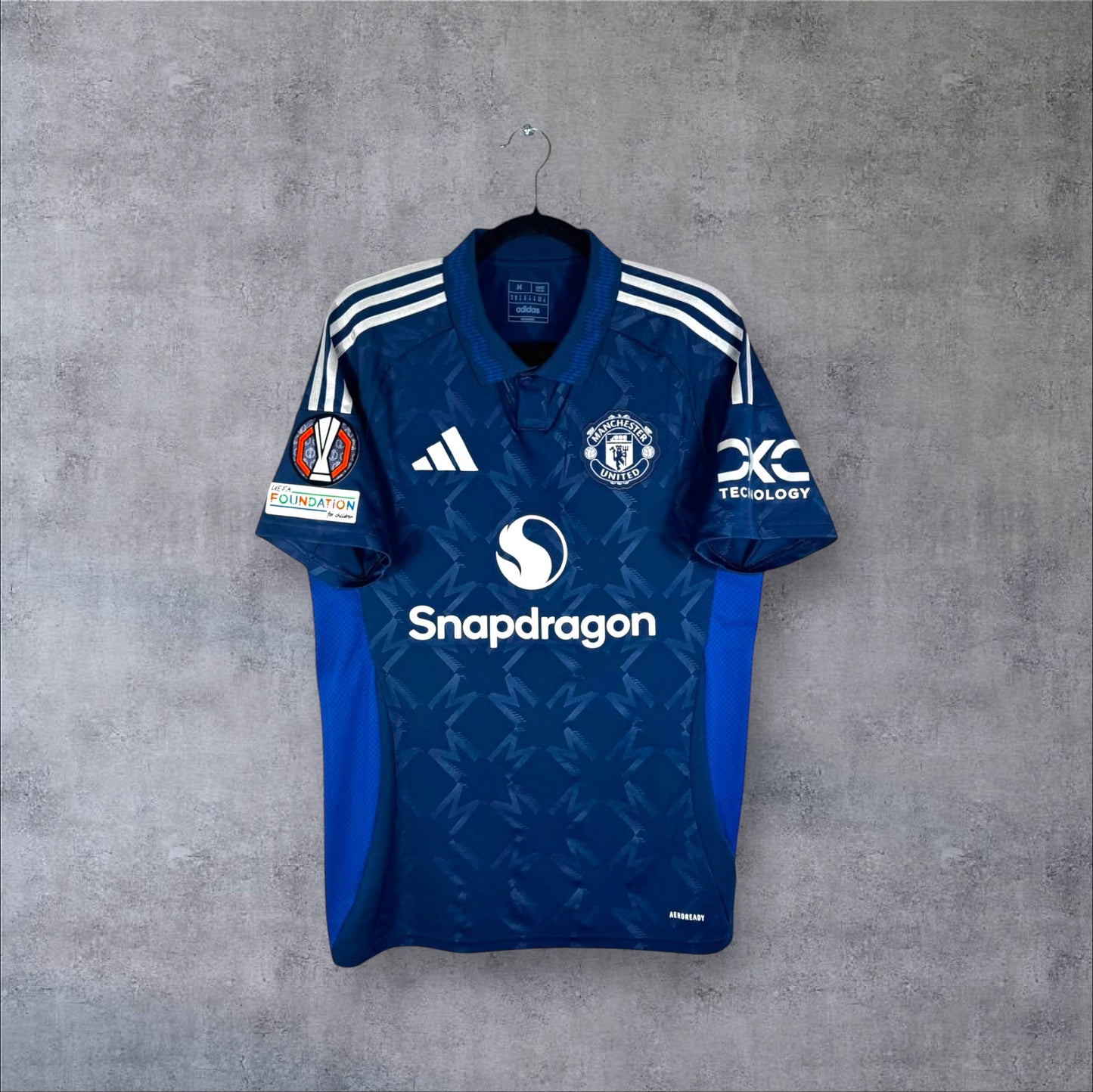 Vue de face du maillot Manchester United Away 2024/25 bleu indigo avec col polo, motif monogramme M et nouveau sponsor Snapdragon.