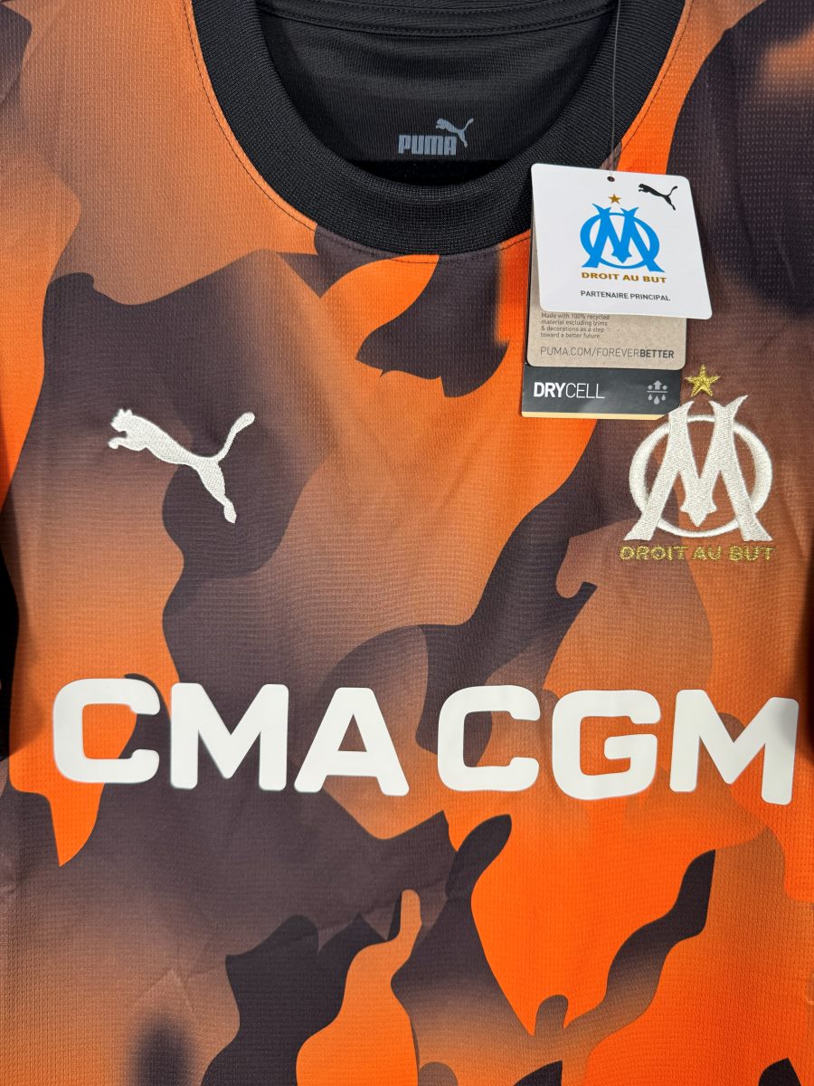 Détail de l'écusson OM brodé doré Droit au But et du sponsor CMA CGM sur le maillot third 2023-2024