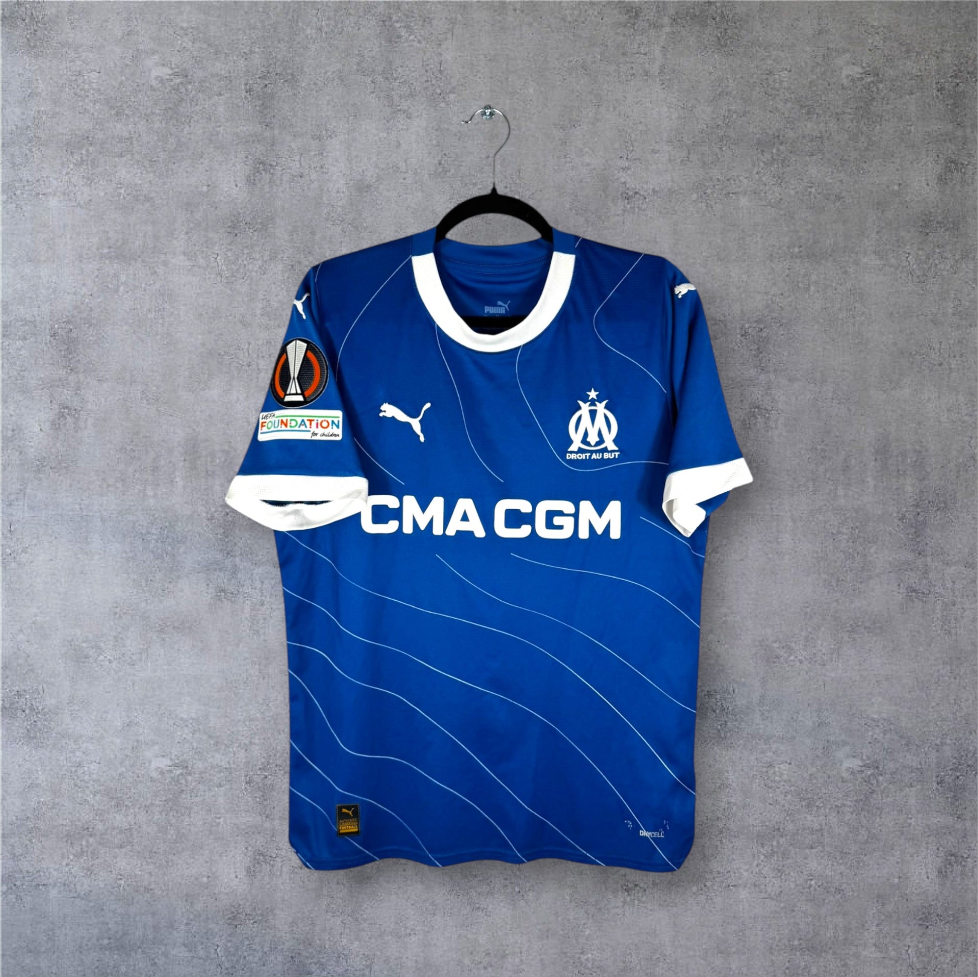 Vue de face du maillot OM Extérieur 2023/24 bleu à lignes blanches ondulées et sponsor CMA CGM.