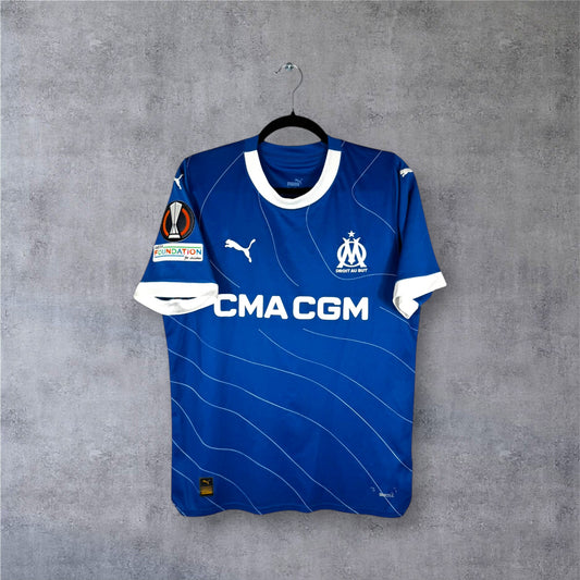 Vue de face du maillot OM Extérieur 2023/24 bleu à lignes blanches ondulées et sponsor CMA CGM.