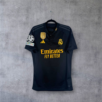 Vue de face du maillot Real Madrid Third 2024 noir avec badge FIFA doré au centre.